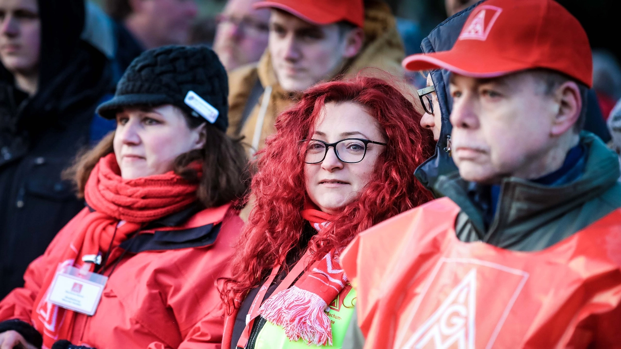 Eine Frau mit roten Locken, dunkler Brille und IG Metall-Schal lächelt in die Kamera.